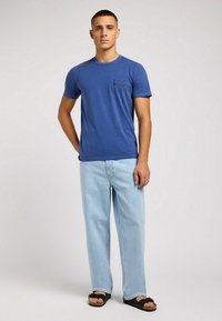 Camiseta de algodón azul con mangas cortas y un escote circular, que presenta un pequeño logo oscuro en el lado izquierdo del pecho. Jeans de corte holgado en azul claro.