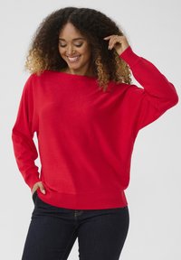 Nainen, jolla on kiharat hiukset, hymyilee, pukeutuneena kirkkaan punaisiin pitkähihaisiin neuleisiin ja tummiin farkkuihin, poseseraa yksi käsi taskussa ja toinen lähellä päätä.