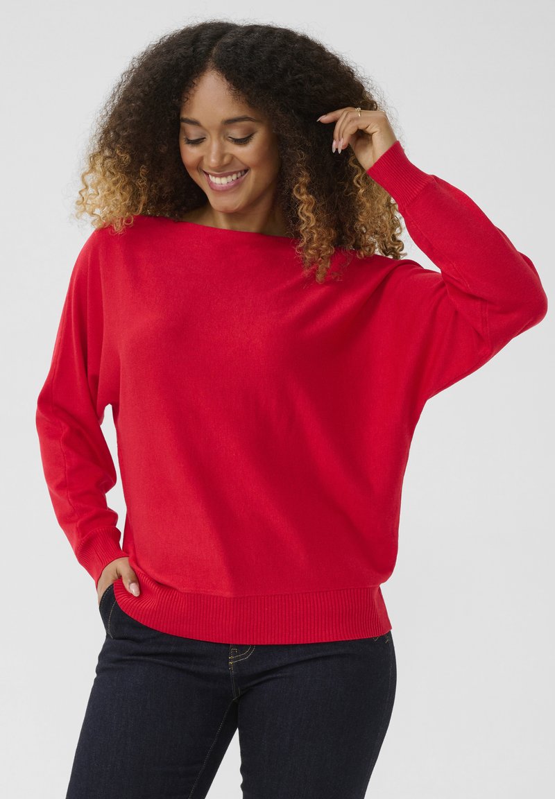 Nainen, jolla on kiharat hiukset, hymyilee, pukeutuneena kirkkaan punaisiin pitkähihaisiin neuleisiin ja tummiin farkkuihin, poseseraa yksi käsi taskussa ja toinen lähellä päätä.