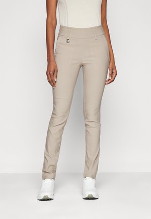 Vrouw draagt beige slim-fit broek met voorzakken, broekriemlussen en witte sneakers, staand tegen een effen lichte achtergrond.