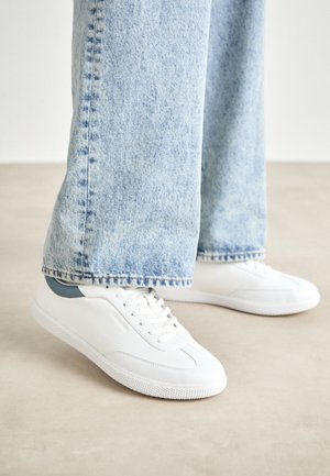 White sneakers with smooth material, lace-up design, textured rubber sole, and a teal accent at the heel, paired with light blue jeans.