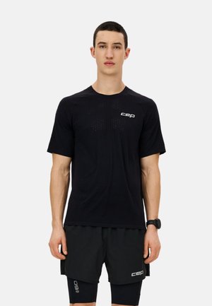 Junger Mann mit kurzen dunklen Haaren, der ein schwarzes CEP-Sportshirt, schwarze Shorts, schwarze Kompressionsleggings und eine schwarze Smartwatch trägt.