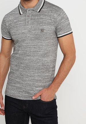 Homme portant un polo gris chiné avec un col et des manches bordés noir et blanc, main dans la poche, sur un fond uni.