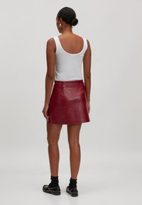 Roter Leder-Minirock mit ausgestellt geschnittenem Saum, Reißverschluss auf der Rückseite und einer glatten Textur. Kombiniert mit einem weißen Trägershirt und schwarzen Socken.