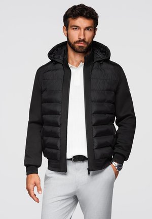 Giubbotto puffo nero con cappuccio, caratterizzato da una texture trapuntata sul petto e maniche lisce. Indossato sopra una polo chiara, abbinato a pantaloni chiari.