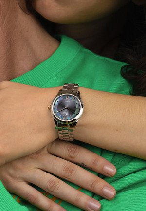 Personne portant une chemise verte avec une montre en métal argenté au poignet, les mains croisées montrant le cadran de la montre avec des chiffres romains et un cadran sombre.