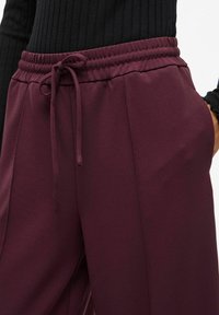 Burgundské tepláky s elastickým pasem, detailem na šňůrku a postranními kapsami, vyrobené z měkké a hladké tkaniny.