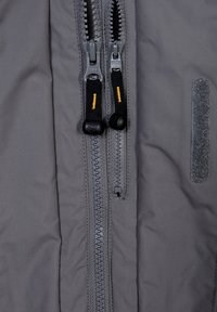 Šedá tkanina bundy se dvěma centrálními zipy. Na jezdcích zipů jsou černé šňůrky se žlutými akcenty. Textura je hladká, vedle je suchý zip.