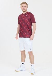 Rød mønstret træningsskjorte med korte ærmer, parret med hvide shorts. Modellen har hvide sko og sokker på, hvilket fremviser en sporty stil.