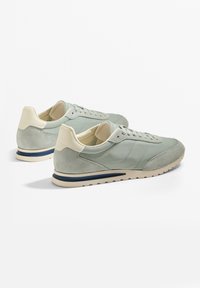 Világoszöld sportcipő velúr és szintetikus felsőrésszel, krém színű részletekkel és tengerészkék csíkkal a talpon, lekerekített orral és fűzőzárral.