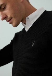 Schwarzer Strickpullover mit V-Ausschnitt, der ein strukturiertes Design und ein kleines gesticktes Logo auf der Brust zeigt. Darunter ein weißes Hemd mit Knöpfen.