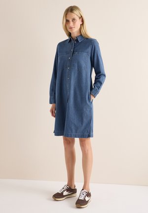 Vrouw draagt een blauwe denim jurk met lange mouwen en voorzakken, gecombineerd met bruine en witte sneakers, staand tegen een neutrale achtergrond.