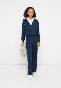 Survêtement bleu marine avec des rayures blanches sur les côtés, fabriqué dans un matériau léger. Comprend une veste zippée et un pantalon ample. Baskets blanches et casquette beige.
