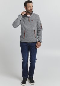 Pulover gri din fleece cu guler cu fermoar pe jumătate, fermoare portocalii și buzunare laterale, asociat cu blugi închiși și adidași negri.