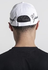 Biely baseballový klobúk s čiernou výšivkou s nápisom "Gianni" na zadnej strane. Má nastaviteľný popruh a je viditeľný zozadu.