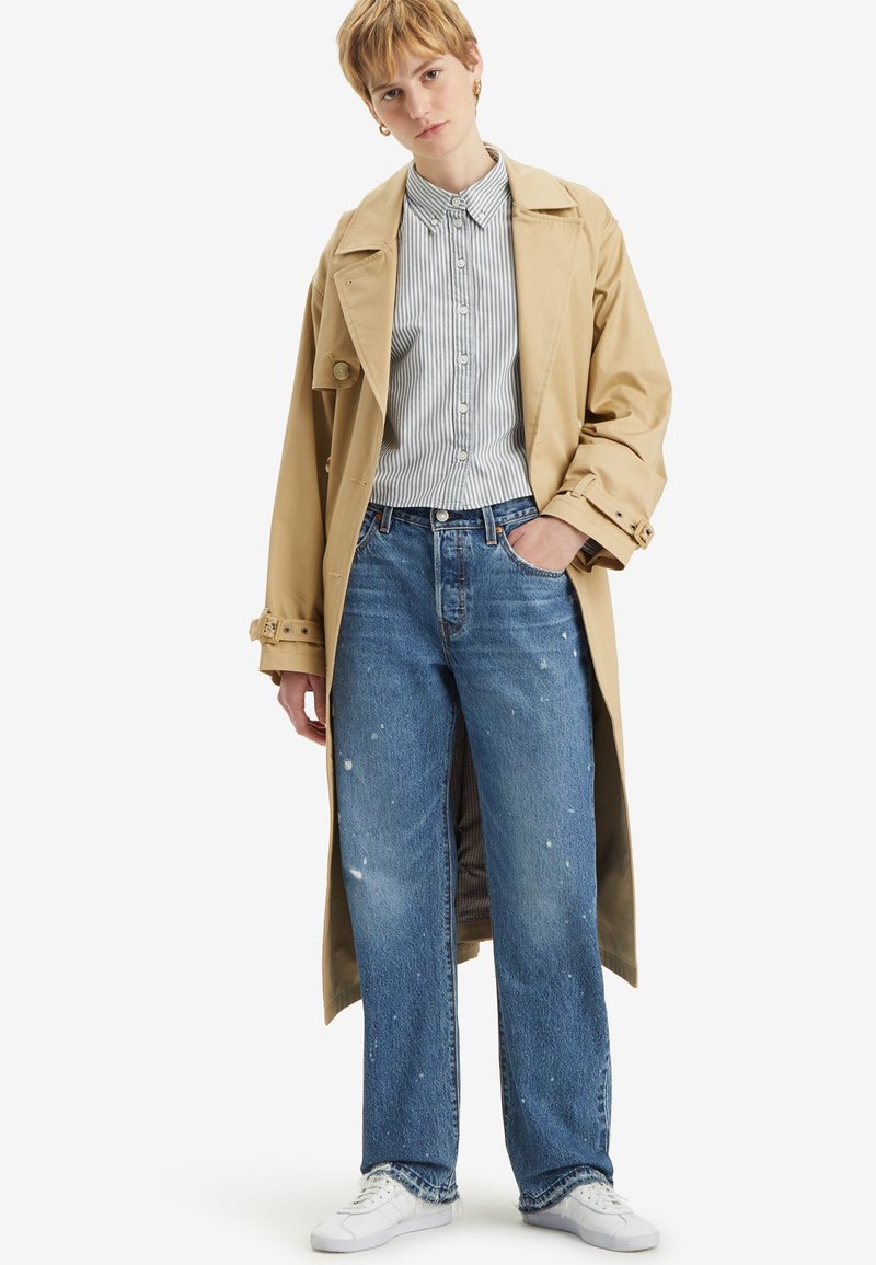 Persona che indossa un trench coat beige, una camicia a righe con bottoni, jeans blu strappati e sneakers bianche, in piedi con una mano in tasca.