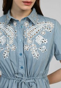Blouse rayée bleu et blanc avec une broderie blanche complexe et des détails en perles sur le col, dotée d'une taille cintrée et de boutons.