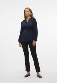 Blusa azul marino con mangas largas y abullonadas, cierre frontal de botones y cinturón atado en la cintura. Combinada con pantalones negros ajustados y zapatos planos.