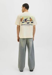 Cream-colored cotton t-shirt featuring a graphic of colorful chairs on the back, paired with loose-fitting light blue jeans.