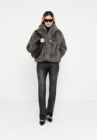Faux fur grå jakke med høj krave, to knapper, matchet med sorte tætsiddende bukser og spidse sorte sko, mens der holdes en sort håndtaske.