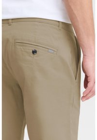 Beige Hosen mit glatter Textur, versehen mit einer Gesäßtasche, die mit einem schwarzen Knopf gesichert ist, und einem kleinen gestreiften Akzent über der Tasche.
