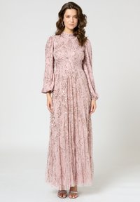 Langes pinkes Kleid mit langen Ärmeln, verziert mit Pailletten in wirbelnden Mustern; durchsichtiger Stoff, fließende Silhouette, ergänzt durch silberne Sandalen.