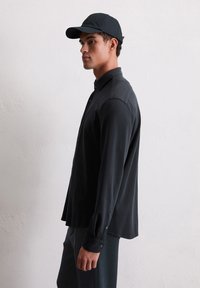 Man wearing all-black outfit including a black cap, long-sleeve buttoned shirt, and pants standing against a plain light wall.