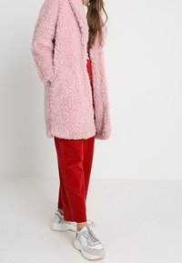 Cappotto fluffy pink con finitura testurizzata, abbinato a pantaloni di velluto a coste rossi e sneakers bianche chunky, presentato in un outfit casual e alla moda.