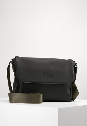 Sac bandoulière noir avec fermeture à rabat, logo "A|X" embossé, et bandoulière réglable vert olive sur une surface blanche.