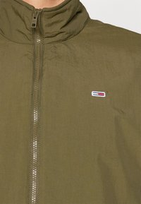 Chaqueta ligera verde oliva con un alto cuello y un cierre completo, que presenta un pequeño logo bordado en el pecho en rojo, blanco y azul.