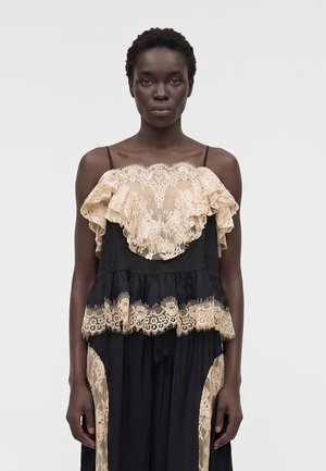 Μαύρη γυναίκα που φοράει μαύρο φόρεμα με μπεζ δαντελένιες βολάν στο στήθος, τη μέση και κατά μήκος των πλευρών σε απλό φόντο.