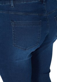 Jean en denim bleu foncé et ajusté montrant les coutures de la poche arrière et de la ceinture sur le côté droit de la partie inférieure du corps.