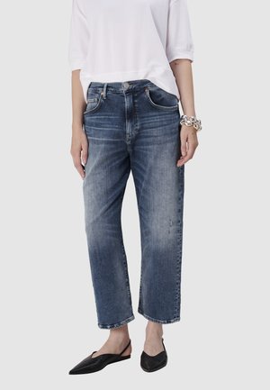 Jean en denim de couleur bleu moyen, avec une coupe décontractée, cinq poches, un léger délavage et une jambe droite. Porté avec des ballerines noires.