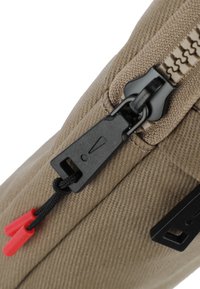 Dettaglio di una borsa in tessuto beige con una zip nera in plastica e accenti rossi in tessuto. La trama superficiale è rigata, mostrando le cuciture lungo le giunture.