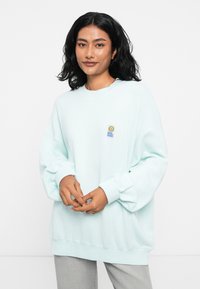 Mėtinės žalios spalvos per didelis džemperis, pagamintas iš fliso, su raštuotomis rankogaliais ir apatine dalimi. Ant kairiojo krūtinės ploto yra mažas, spalvingas logotipas.