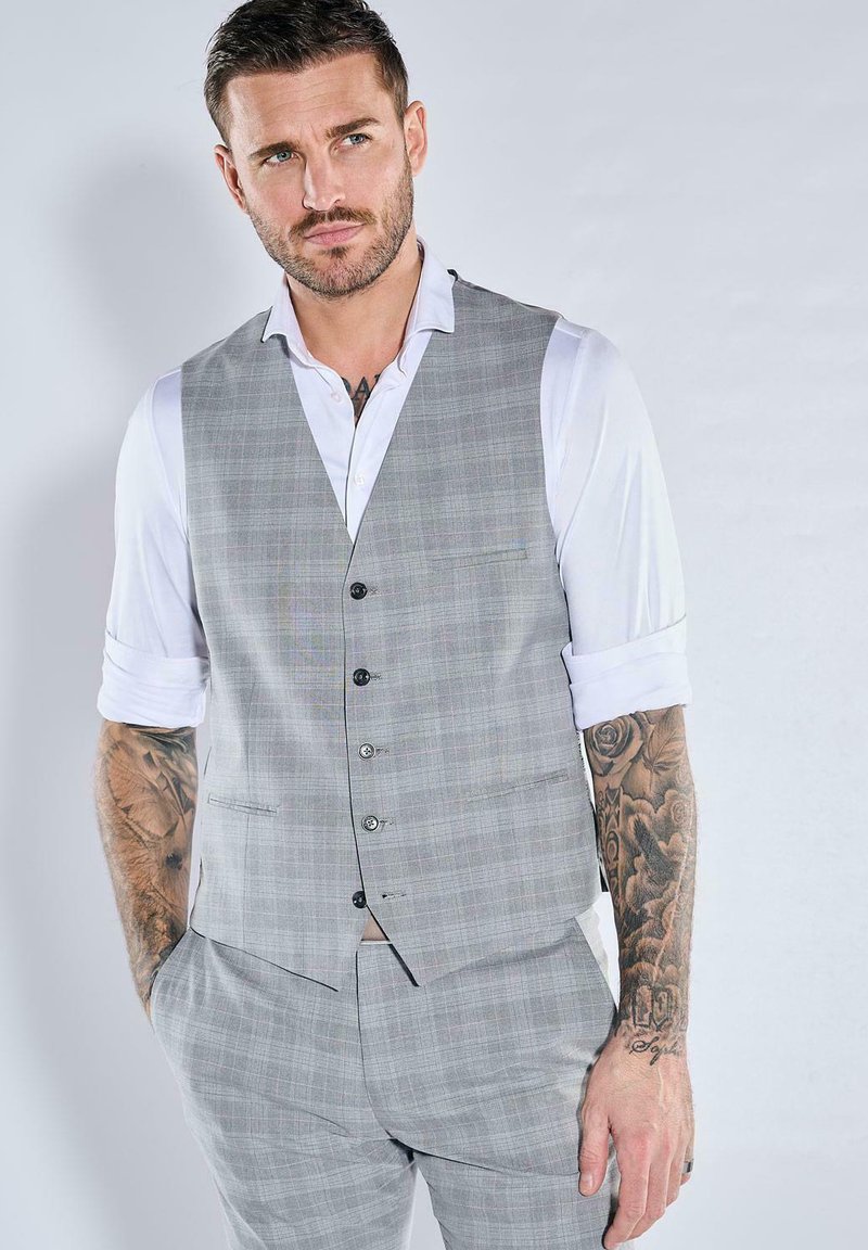 Graue karierte Weste über einem weißen Hemd mit hochgekrempelten Ärmeln, mit schwarzen Knöpfen und Brusttaschen. Tätowierung am linken Arm sichtbar.