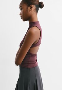 Jonge vrouw met haar in een knot draagt een mouwloze kastanjebruine top met een uitgesneden achterkant en een donker hoge taille rok, staat zijwaarts.