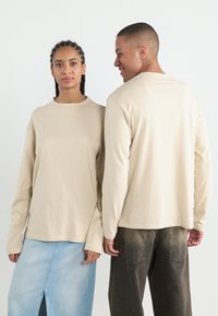 Beige long-sleeve t-shirts on two figures, featuring a crew neck and relaxed fit, paired with a light blue denim skirt and dark trousers.
