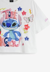 Vit kortärmad croptop med en blå tecknad karaktär med öppen mun, färgglada blommor, en fjäril och texten "TROUBLEMAKER!" i fet stil.