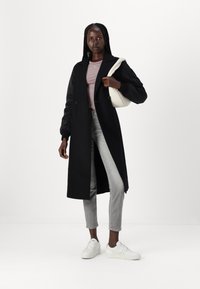 Lange zwarte jas met kraag en knoopdetails, gecombineerd met een gestreepte roze top en grijze jeans. Accessoires zijn onder andere een lichtgekleurde schoudertas.