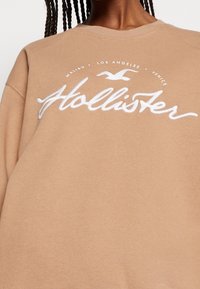Beige sweatshirt i mjukt tyg, med en vit broderad logotyp med en fågel och text som anger platser. Rund halsringning, avslappnad passform.