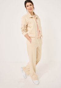 Veste en coton beige avec col et fermeture à boutons, associée à un pantalon beige. En dessous, une chemise blanche à rayures orange. Chaussures blanches.
