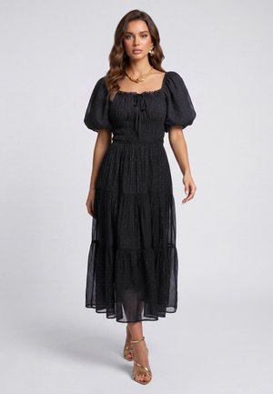 Femme portant une robe midi noire à manches bouffantes et petits pois, associée à des sandales dorées, un collier et des boucles d'oreilles sur un fond gris uni.
