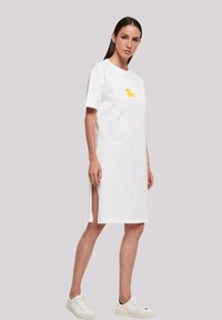 Weißes Baumwollkleid mit kurzen Ärmeln, seitlichen Schlitzen und einem gelben Gummiente-Motiv auf der Vorderseite. Getragen mit weißen Turnschuhen.