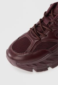 Burgundské športové tenisky s kombináciou sieťoviny a kože, textúrovanými panelmi, zaoblenou špičkou a hrubou, vzorovanou podrážkou.