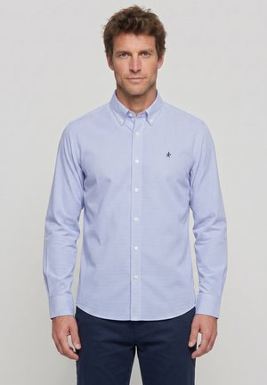 Hombre de pie frente a un fondo liso, vistiendo una camisa de cuadros azul claro abotonada y pantalones oscuros, mirando hacia adelante con expresión neutral.