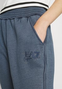 Blue joggers made of soft fabric featuring a ribbed waistband, side pockets, and an embroidered EA7 logo with decorative studs.
