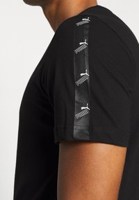 Camiseta negra de manga corta con una tela suave, con una franaja lateral negra brillante y logotipos de Puma en blanco repetidos en el hombro.