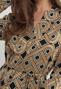 Langärmliges Kleid mit einem schwarz-goldenen geometrischen Muster, V-Ausschnitt und gebundenem Bund für eine figurbetonte Passform. Weicher Stoff.