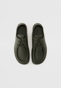 Sötétzöld bőr slip-on cipők, lekerekített orral, díszes hurok záródással és texturált talppal. Simító felület.